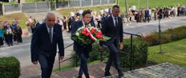 Trzech mężczyzn w formalnych strojach niesie kompozycję kwiatową, wchodząc po schodach na uroczystość upamiętniającą, z tłumem ludzi w tle.