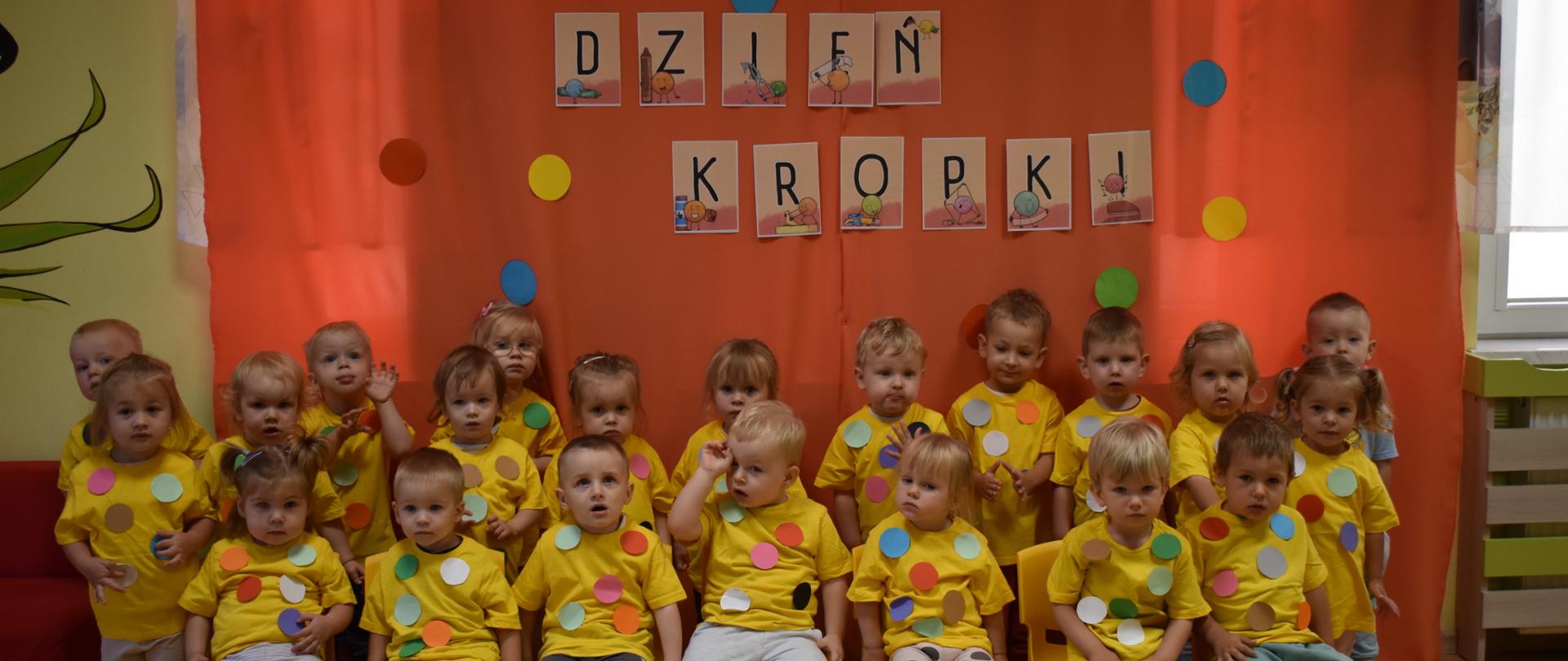 Na zdjęciu widać dzieci z grupy Tygryski. Wszystkie są ubrane w żółte koszulki z naklejonymi kolorowymi kropkami. W tle pomarańczowa ścianka z napisem dzień kropki.