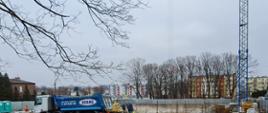 Zdjęcie przedstawia postęp aktualnych prac budowlanych, pracownicy przygotowują zbrojenia, samochody ciężarowe przywożą materiały na plac budowy.