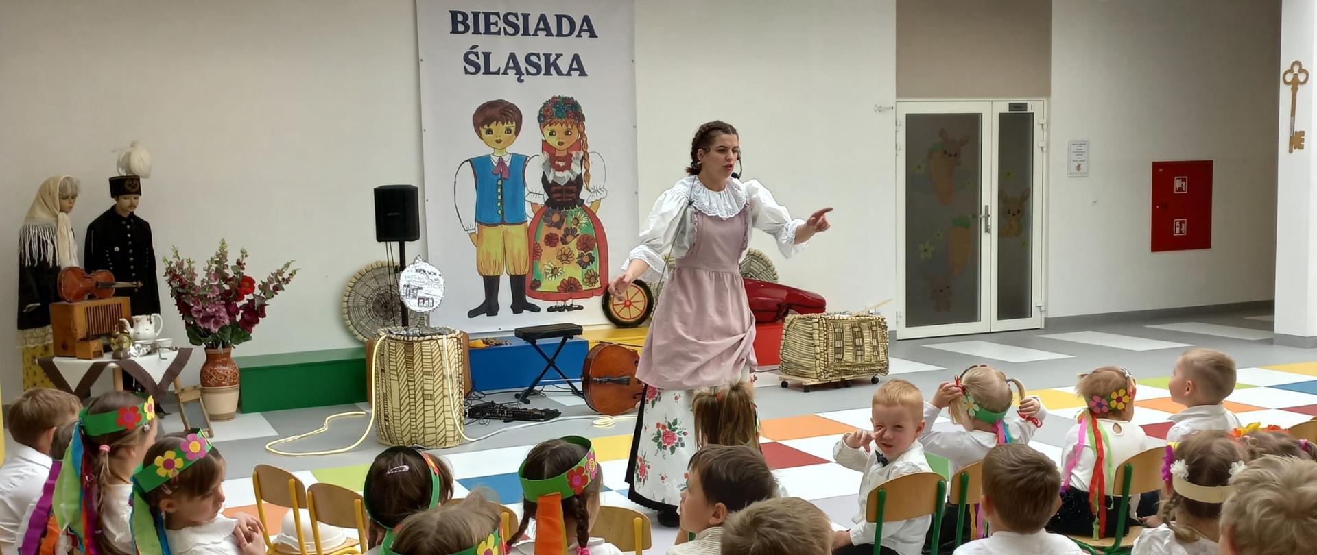 Kobieta w stroju śląskim stoi przed grupą dzieci siedzących na krzesełkach