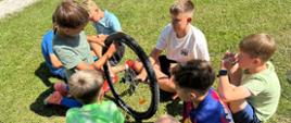 Dzieci siedzą na trawie i uczą się jak wymienić dętkę w rowerze.