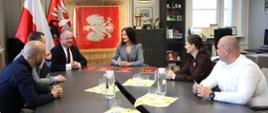 Na zdjęciu widoczna jest grupa sześciu osób siedzących przy dużym, ciemnym stole konferencyjnym podczas oficjalnego spotkania. W centralnej części tła, za uczestnikami, znajdują się polskie symbole narodowe oraz lokalne, w tym flaga Polski i godło z orłem. Na stole rozstawione są szklanki, karafki z wodą oraz czerwone teczki z dokumentami.