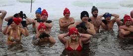 Zdjęcie przedstawia liczną grupę osób biorących udział w VI Karnawałowym Zlocie Morsów w Zambrowie. Uczestnicy znajdują się w lodowatej wodzie zambrowskiego Zalewu Miejskiego. W tle widać zaśnieżony brzeg, drzewa oraz zabudowania miejskie pod zachmurzonym niebem. Morsujący są ubrani w stroje kąpielowe oraz kolorowe czapki (przeważają czerwone), a niektórzy mają na sobie karnawałowe akcesoria, jak naszyjniki z kwiatów. W centrum grupy widoczne są dwie duże flagi: jedna czerwona z logo klubu, a druga błękitna z hasłem: "Zadbaj o zdrowie! Hartuj się z morsami!" Atmosfera jest radosna.