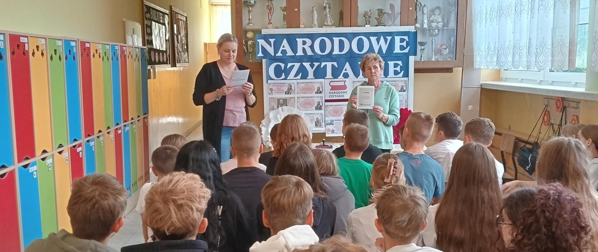 Pani wicedyrektor odczytuje fragment utworu Jana Kochanowskiego, obok stoi bibliotekarka szkolnej biblioteki, za paniami stół nakryty jasnym płótnem i gazetka informacyjna związana z imprezą z napisem "Narodowe Czytanie", na przodzie na małych ławeczkach siedzą słuchający uczniowie