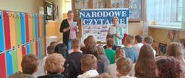 Pani wicedyrektor odczytuje fragment utworu Jana Kochanowskiego, obok stoi bibliotekarka szkolnej biblioteki, za paniami stół nakryty jasnym płótnem i gazetka informacyjna związana z imprezą z napisem "Narodowe Czytanie", na przodzie na małych ławeczkach siedzą słuchający uczniowie