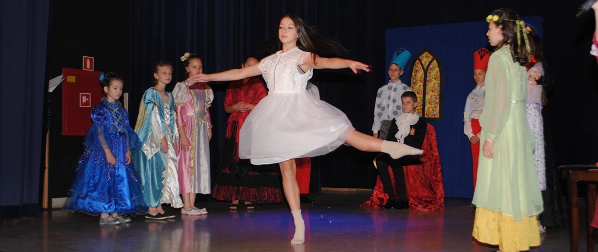 Dziewczynka ubrana w białą sukienkę tańczy na scenie, w tle inne dzieci kolorowo ubrane stoją i się przyglądają 