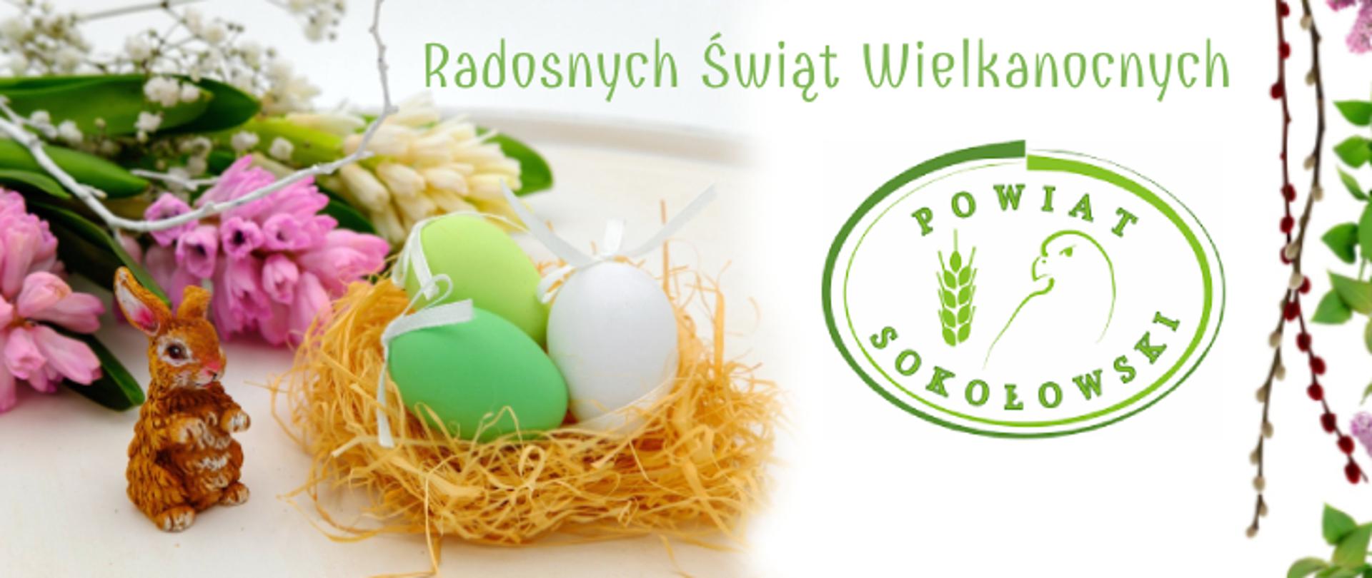 Grafika przedstawia ilustrację wielkanocną. z lewej strony leży w tle bukiet kwiatów w kolorach różu i bieli na pierwszym planie siedzi zajączek z łapkami wzniesionymi ku górze, obok w gniazdku z sianka leżą trzy pisanki zielona, limonkowa i biała z wywiązanymi białymi kokardkami, po prawej stronie jest logo Powiatu Sokołowskiego, obok widać dwie palemki wielkanocne oraz zielone liski i fragmenty różowych kwiatów. na górze grafiki widnieje zielony napis "Radosnych Świąt Wielkanocnych".