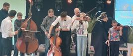 Muzycy na scenie pomagają dzieciom trzymać i próbować grać na instrumentach smyczkowych, takich jak kontrabas, wiolonczela i skrzypce. Występ odbywa się w szkolnej sali gimnastycznej przed siedzącą na podłodze widownią złożoną z uczniów.