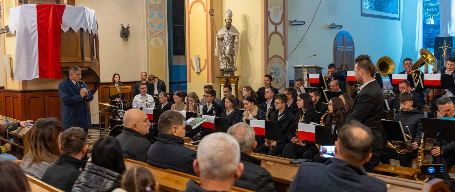 Wnętrze kościoła podczas koncertu patriotycznego. Na pierwszym planie siedzą mieszkańcy, a na prezbiterium Parafialna Orkiestra Dęta z Leńcz w pełnym składzie z biało czerwonymi flagami na pulpitach. Z lewej strony stoi Burmistrz Kalwarii Zebrzydowskiej Tadeusz Stela, który przemawia do zgromadzonych z mikrofonem w dłoni. W tle widać oświetlone wnętrze świątyni, elementy dekoracyjne oraz rzeźbę biskupa.
