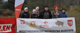 Zawodnicy i zawodniczki z drużyny z trenerem pozują do zdjęcia przy banerze Wojewódzkich Mistrzostwach Młodzików w biegach przełajowych
