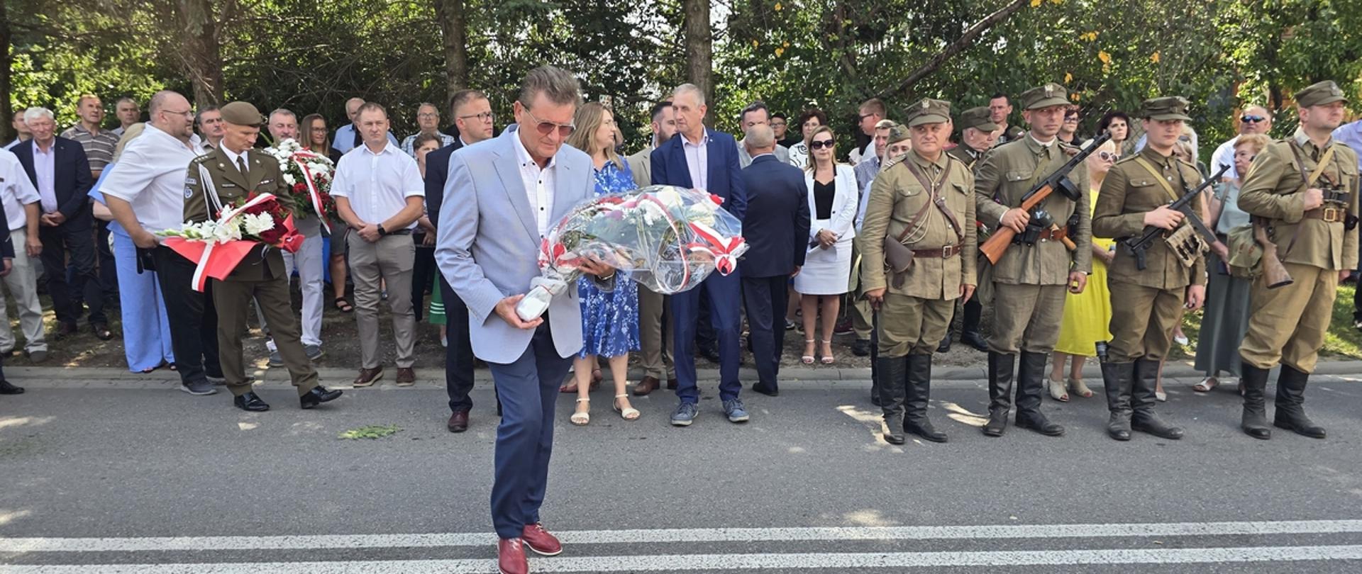 Na fotografii przewodniczący rady Powiatu Sokołowskiego idzie w kierunku pomnika, aby złożyć biało czerwone kwiaty z szarfami patriotycznymi. Za nim czekają inne delegacje, aby złożyć kwiaty. Z prawej strony grupa mężczyzn ubrana w wojskowe zielone stroje, trzymają broń, na głowie kaszkiety.