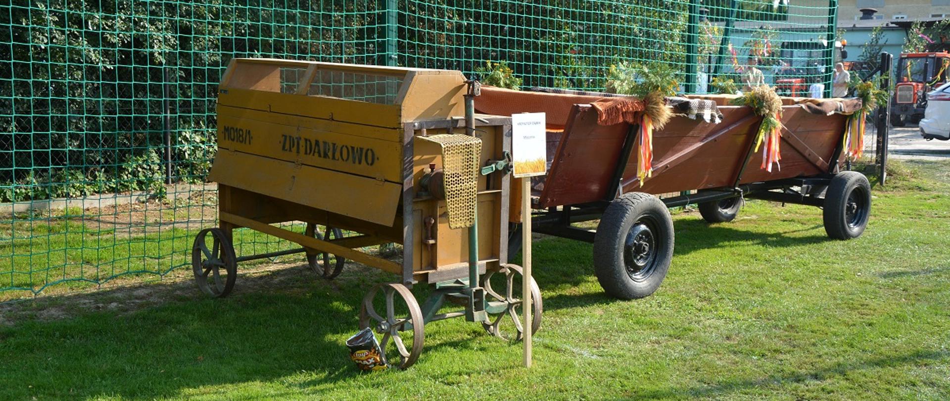 zabytkowa żółta maszyna rolnicza, obok niej stoi drewniany wóz przybrany wstążkami kolorowymi 