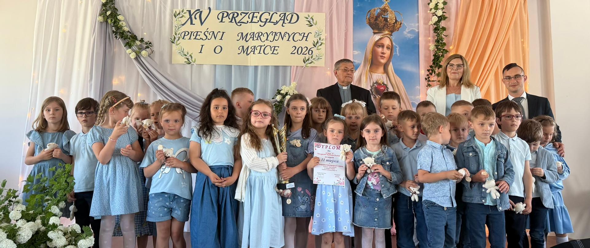 Liczna grupa przedszkolaków w błękitnych strojach pozuje na scenie z pamiątkowym dyplomem za II miejsce oraz złotym pucharem. W tle, za dziećmi, stoją dorośli opiekunowie – kobieta oraz dwaj mężczyźni (w tym jeden w koloratce).