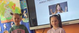 Dwie dziewczynki stoją przed monitorem interaktywnym, na którym wyświetlona jest scenka związana z agresją