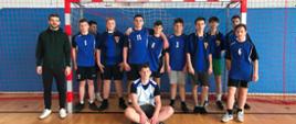 Chłopcy w strojach sportowych, niebieskich koszulkach stoją obok siebie na tle bramki. Obok nich stoi trener