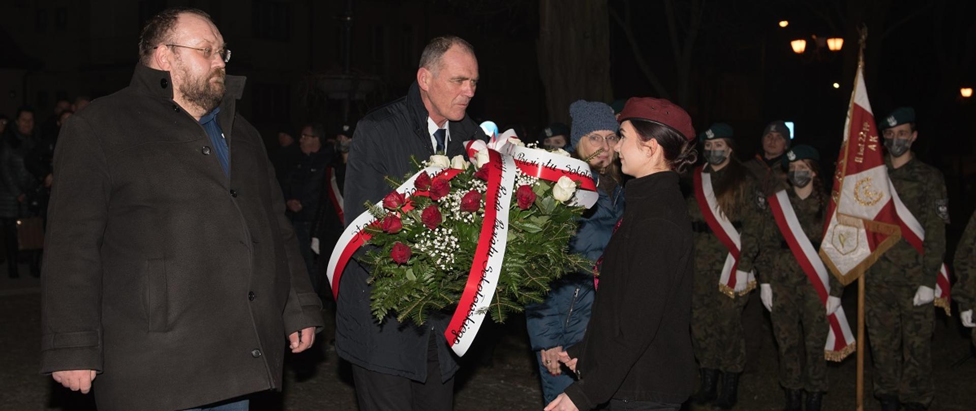 Przewodniczący Rady Powiatu Sokołowskiego Jacek Odziemczyk wraz z wicestarostą Ryszardem Domańskim oraz dyrektor Zespołu Szkół Nr 1 Małgorzatą Baranowską przekazują do rąk harcerki Hufca ZHP w Sokołowie Podlaskim celem złożenia pod pomnikiem. W głębi widoczny sztandar Zespołu Szkół Nr 1 oraz uczniowie klas mundurowych. Fotografia zrobiona w godzinach wieczornych.