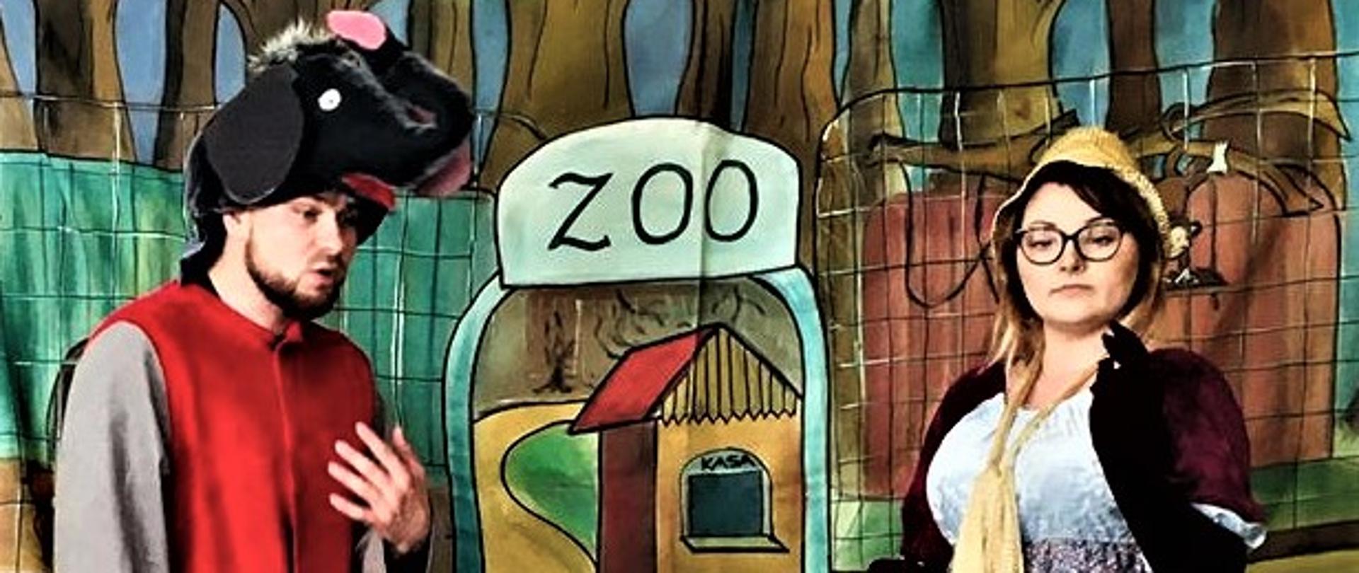Mężczyzna w czerwonym serdaku z pluszową głową konia na głowie, kobieta w okularach . Za nimi namalowane ogrodzenie z bramą z napisem ZOO