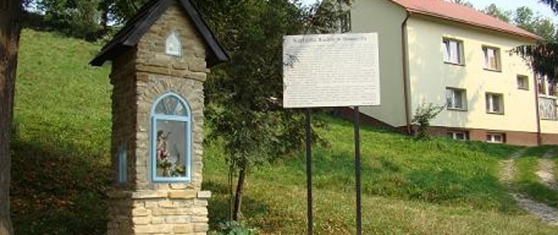 odnowiona kapliczka z tablicą informacyjną wykonana z kamienia przeszklenie z ramą w kolorze niebieskim schodki wejściowe w tle łąka i budynek