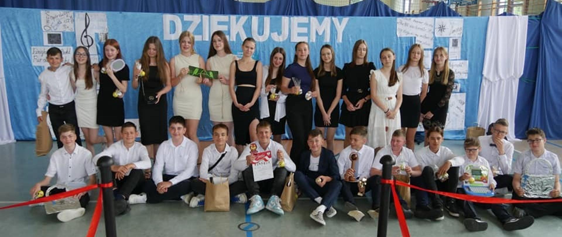 W budynku hali sportowej szkoły podstawowej na tle niebieskiej dekoracji z napisem Dziękujemy ustawieni są absolwenci klas ósmych, którzy w rękach trzymają drobne upominki od sowich młodszych kolegów z klasy 7.
