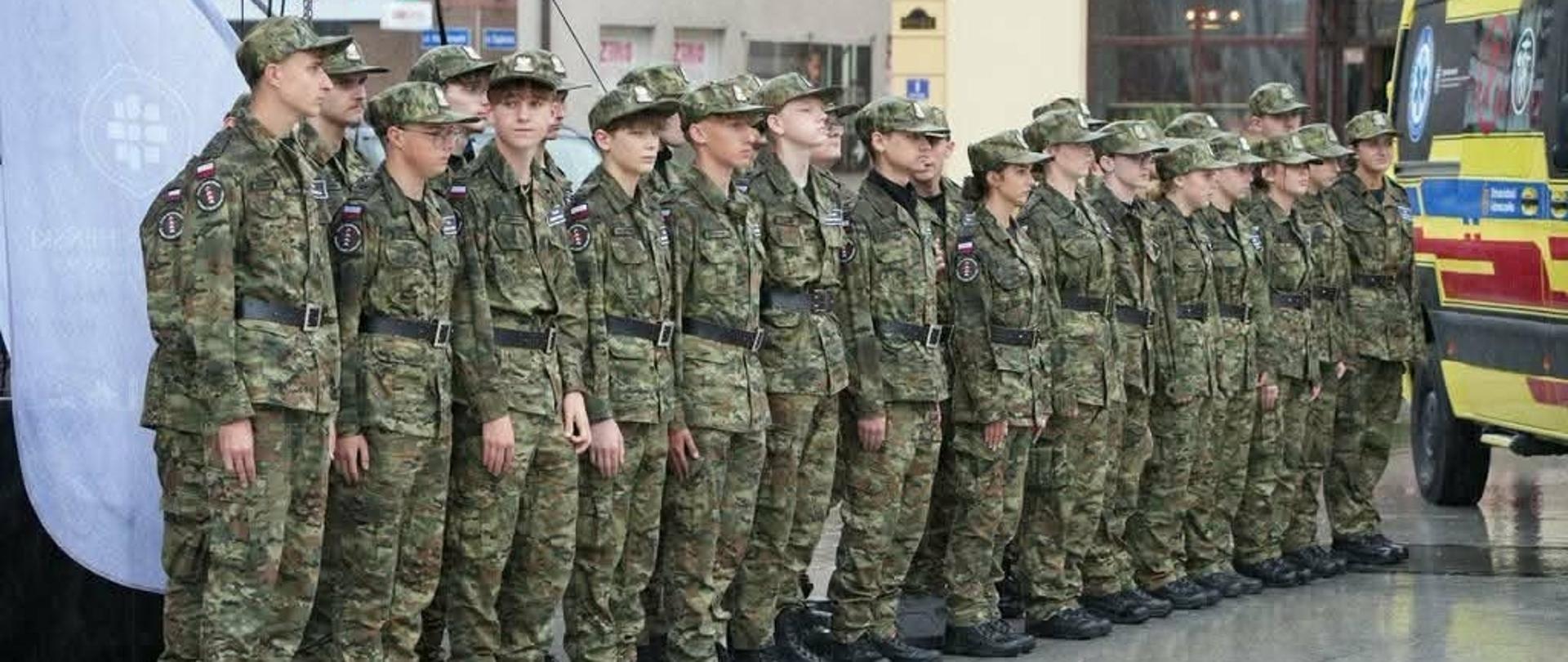 Grupa młodzieży w mundurach polowych stoi w równym szeregu na mokrym placu podczas oficjalnego zgromadzenia. Obok oddziału parkuje żółta karetka pogotowia, a w tle widać miejskie zabudowania z szyldem apteki.