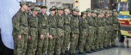 Grupa młodzieży w mundurach polowych stoi w równym szeregu na mokrym placu podczas oficjalnego zgromadzenia. Obok oddziału parkuje żółta karetka pogotowia, a w tle widać miejskie zabudowania z szyldem apteki.
