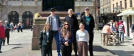 Zdjęcie przedstawia sześcioosobową grupę młodzieży pozującą na słonecznym miejskim placu u stóp pomnika. W tle wznosi się okazały, zabytkowy gmach o bogato zdobionej fasadzie i charakterystycznym, spadzistym dachu.