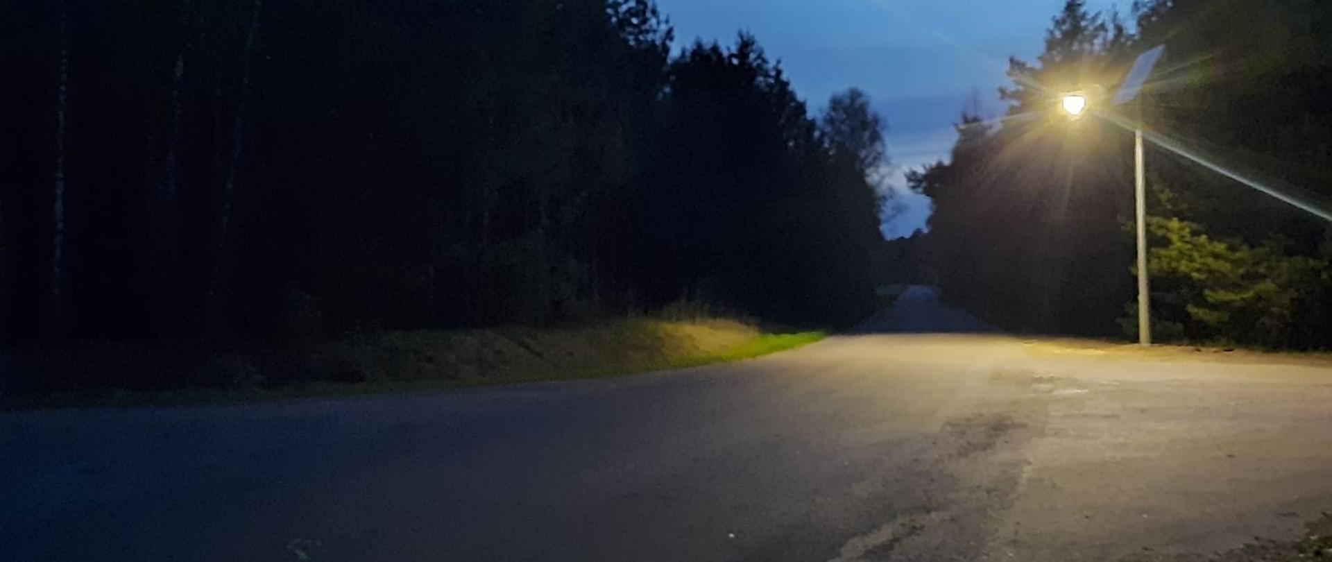 Zdjęcie przedstawia skrzyżowanie drogi powiatowej numer 3223 i drogi gminnej w gminie Jednorożec. Na skrzyżowaniu widnieje jaśniejąca lampa solarna, oświetlająca niebezpieczne po zajściu słońca miejsce na drodze. Zdjęcie wykonano wieczorem.