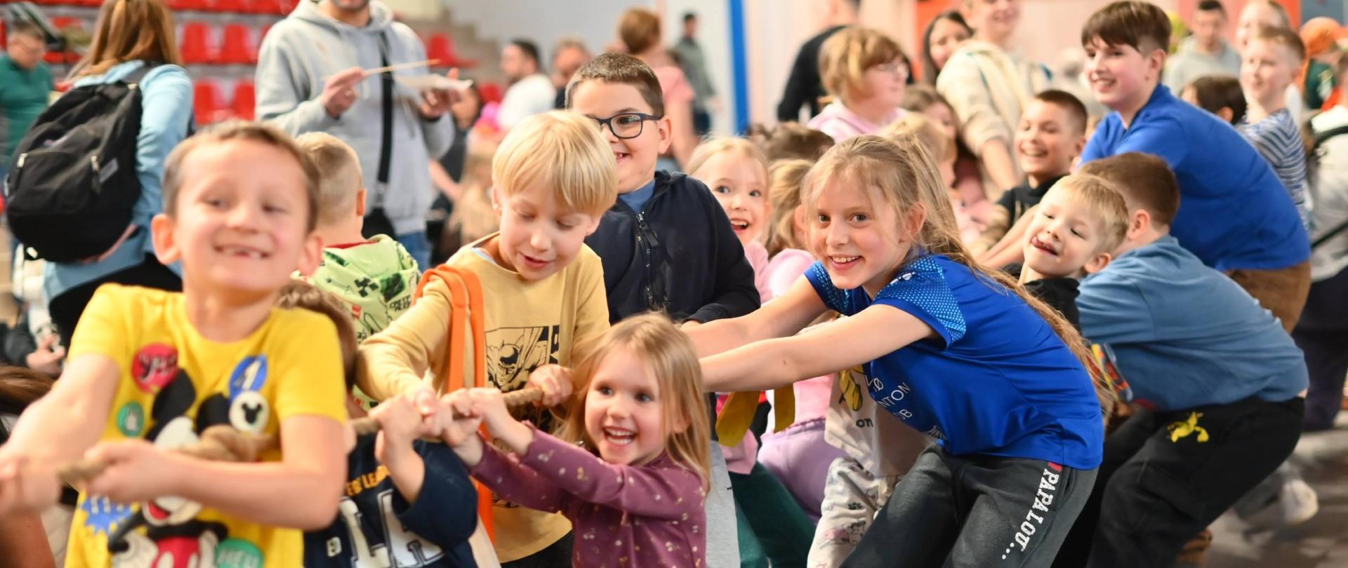 Grupa dzieci bierze udział w zabawie zespołowej, wspólnie ciągnąc linę podczas animacji w hali sportowej; wokół stoją inni uczestnicy i opiekunowie.