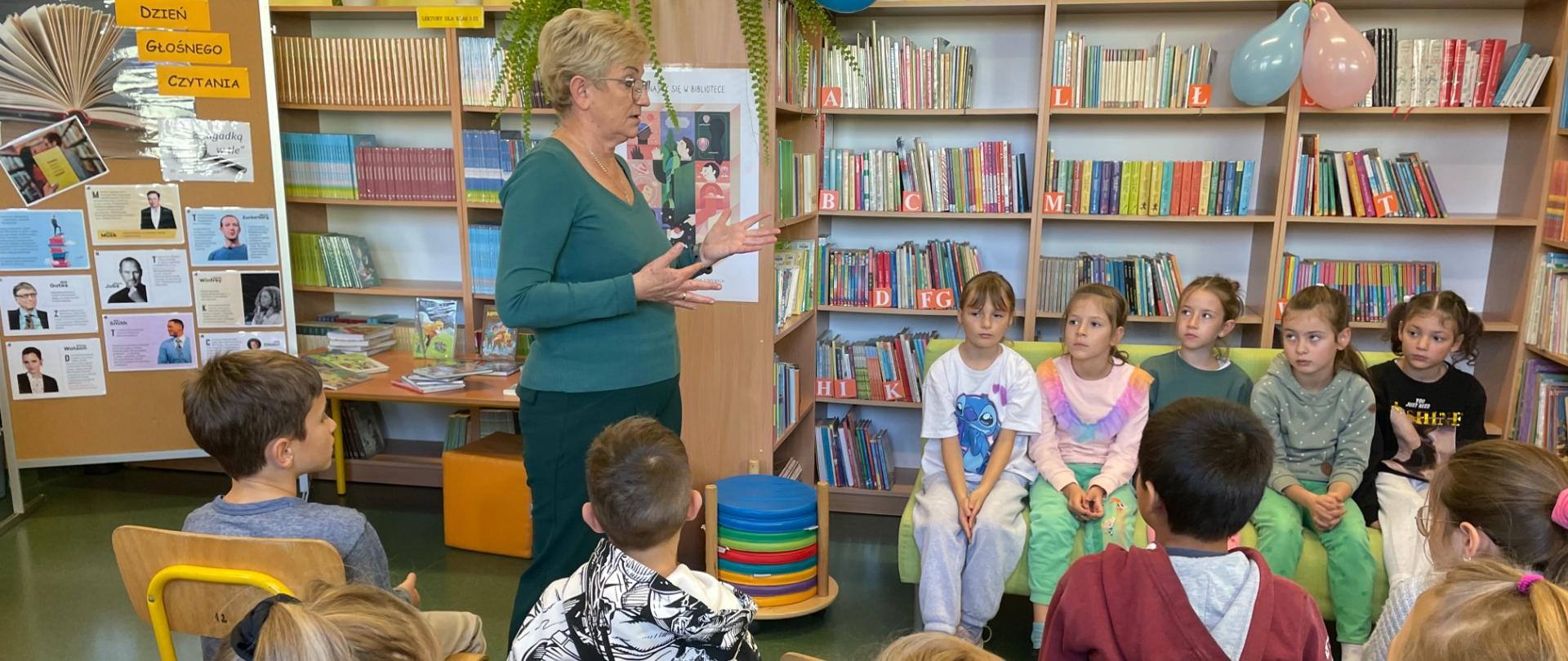 Bibliotekarka stoki przed grupą dzieci siedzących na krzesełkach. W tle półki z książkami.