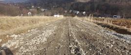 Na środku zdjęcia widoczna odbudowana droga gruntowa po lewej i prawej widoczne fosy oraz łąki na dalszym planie las oraz zabudowania okres zimowy brak śniegu 