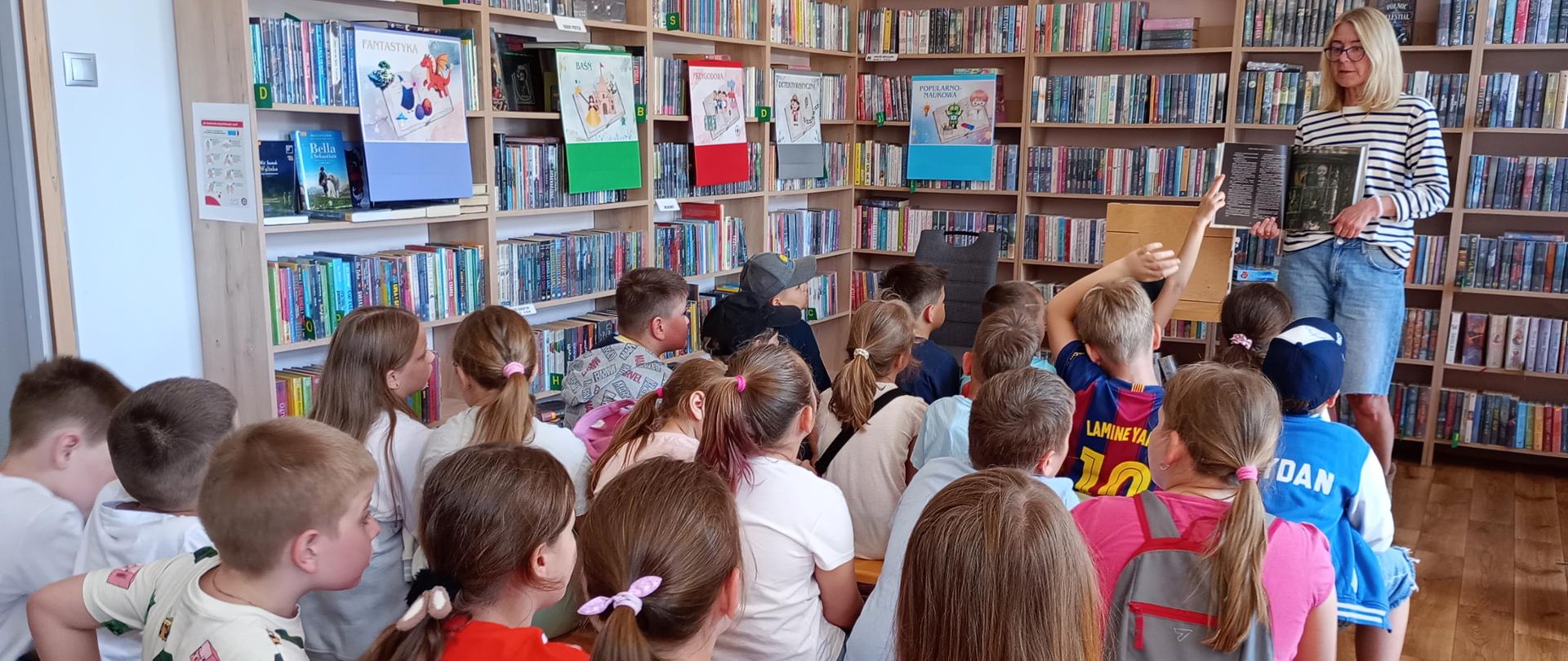 Na zdjęciu widać dzieci od tyłu siedzące ławeczkach wśród regałów i patrzące na bibliotekarkę prezentującą im książkę. Jedno z dzieci zgłasza się by coś powiedzieć. Na regałach z książkami wiszą kolorowe plakaty.