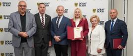 Osoby stoją na tle ścianki do zdjęć i trzymają kartke papieru