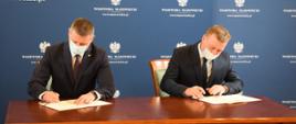 Dwoje mężczyzn siedzących przy stole ubranych w garnitury i białe maseczki na twarzy podpisuje dokumenty