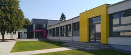 Jednopoziomowy budynek, z płaskim dachem, ściany szare z żółtymi i różowymi wstawkami wokół wejść.
