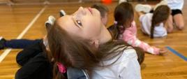 Dziewczynka na podłodze sali gimnastycznej popbiera się przed sobą rękami, odchyla głowę do tyłu i dostaje do niej palcami stóp, w tle widać inne ćwiczące dzieci