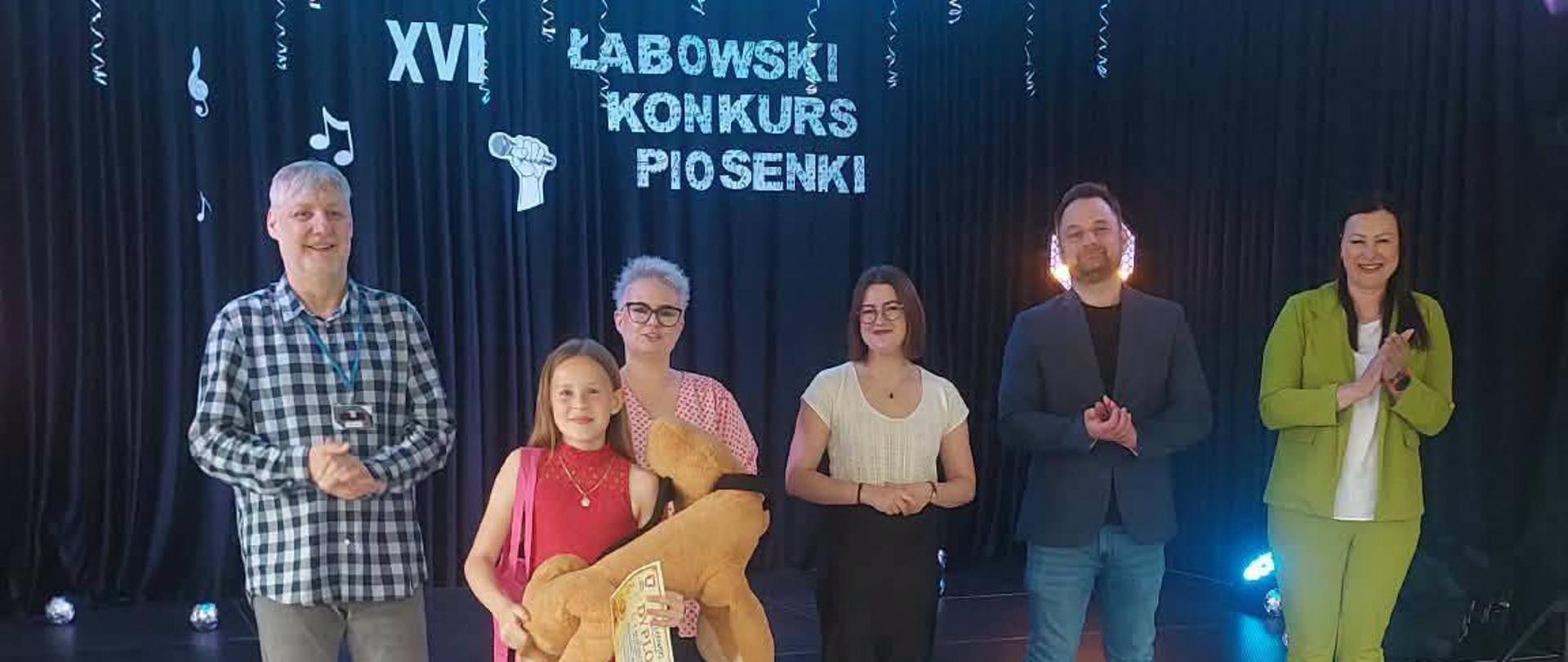 Karolina trzyma dużego brązowego misia, którego otrzymała jako nagrodę, za nią stoi jury konkursu