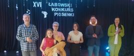 Karolina trzyma dużego brązowego misia, którego otrzymała jako nagrodę, za nią stoi jury konkursu