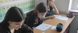 Trzy uczennice siedzą przy stolikach i czytają treść leżących przed nimi kartek.