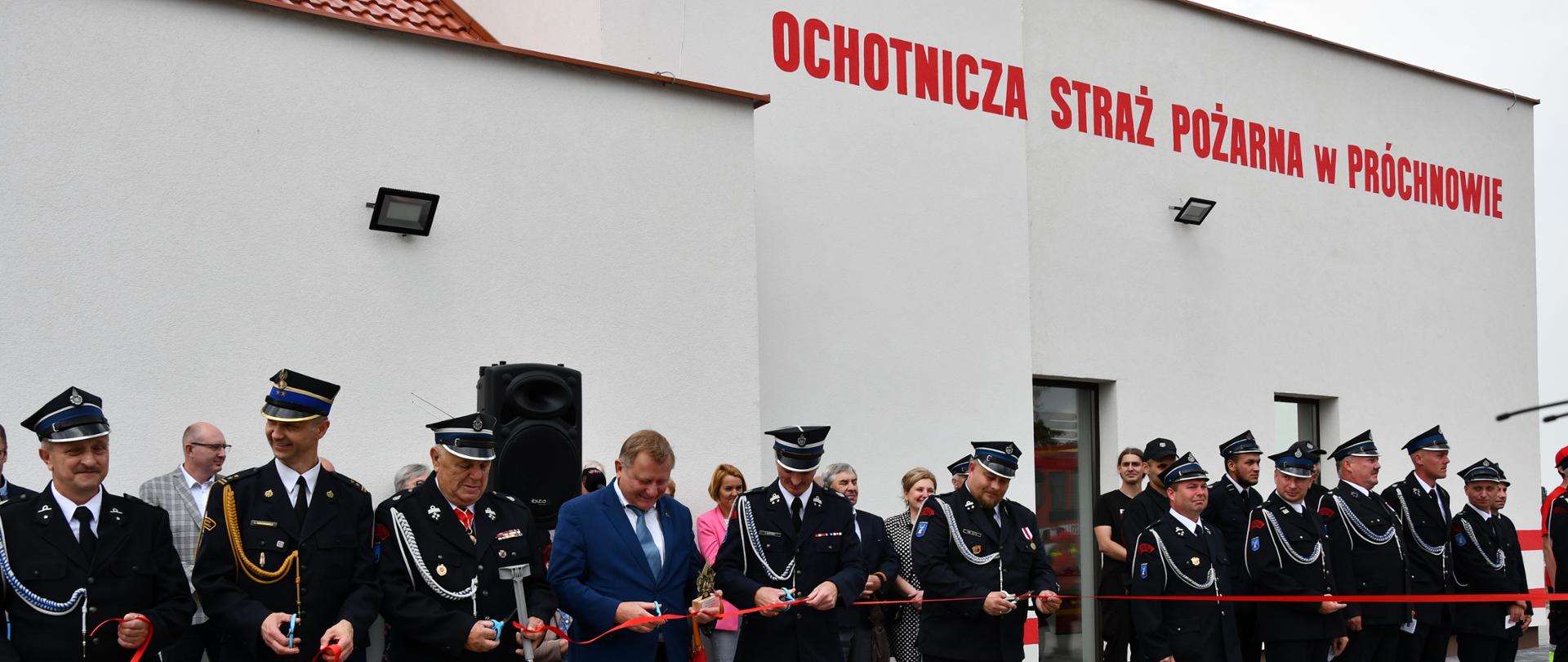 Osoby w mundurach straży pożarnej i osoba w granatowym garniturze przecinają wstęgę na tle biało-szarego budynku z czerwonym napisem Ochotnicza Straż Pożarna w Próchnowie