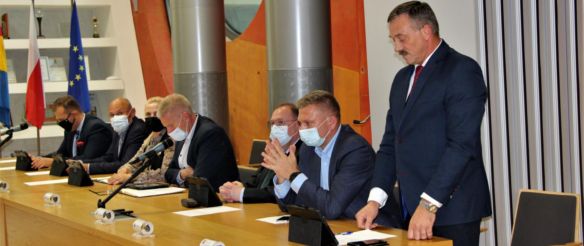 Stojący mężczyzna w garniturze po lewej stronie siedzący mężczyźni i kobieta. W tle flagi od lewej strony flaga powiatu polkowickiego, flaga polski oraz unijna. 