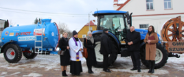 Zdjęcie przedstawia grupę sześciu osób stojących obok niebieskiego ciągnika rolniczego. Do traktora podpięta jest duża, niebieska cysterna z napisem "WODA DO PICIA". Starosta Zambrowski siedzi wewnątrz pojazdu. Wśród osób znajduje się duchowny w szatach liturgicznych oraz mężczyźni i kobiety w zimowych płaszczach. W tle widoczny jest fragment drewnianego koła z napisem "Gmina Szumowo".