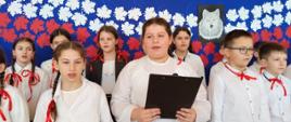 Uczniowie klasy piątej ubrani na galowo, dziewczynki z czerwonymi kokardkami we włosach, chłopcy z czerwonymi kokardkami pod szyją, występują w akademii, za nimi granatowa dekoracja z biało-czerwonymi listkami i wizerunkiem wilka na środku