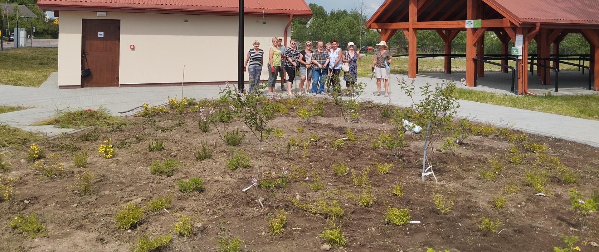 Uczestnicy Klubu Senior+, pozujący do grupowego zdjęcia przed skwerem zieleni na targowicy miejskiej