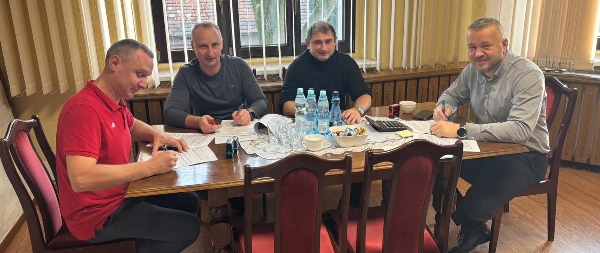 Czterech mężczyzn podpisuje dokumenty siedząc przy ciemnym stole.