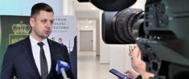 Zdjęcie przedstawia jedną osobę stojącą wewnątrz budynku i udzielającą wywiadu dla telewizji. Osoba wypowiada się do mikrofonu, który trzyma w ręce inna osoba.
