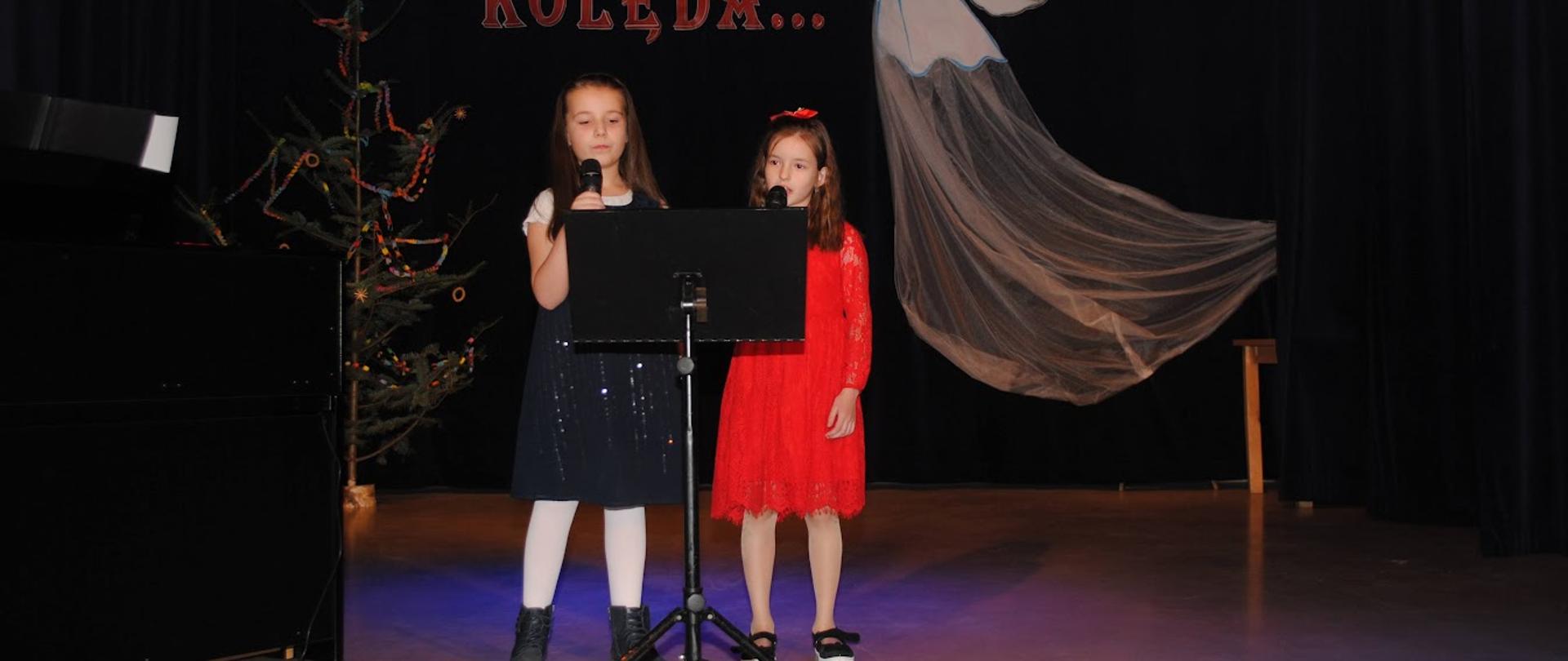 dwie dziewczynki ubrane w ciemną i czerwoną sukienkę stoją na scenie z mikrofonami 