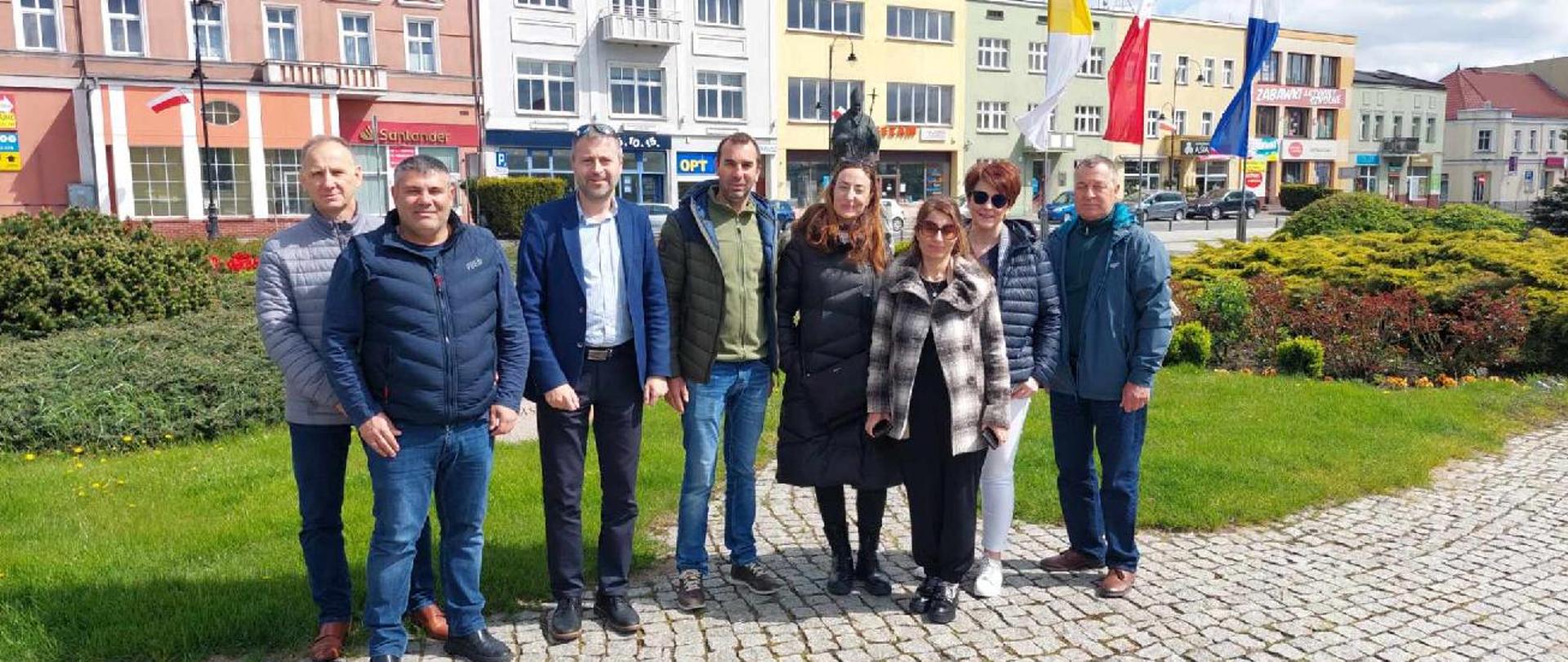 8 osób stojących na kostce brukowej na tle budynków wokół rynku