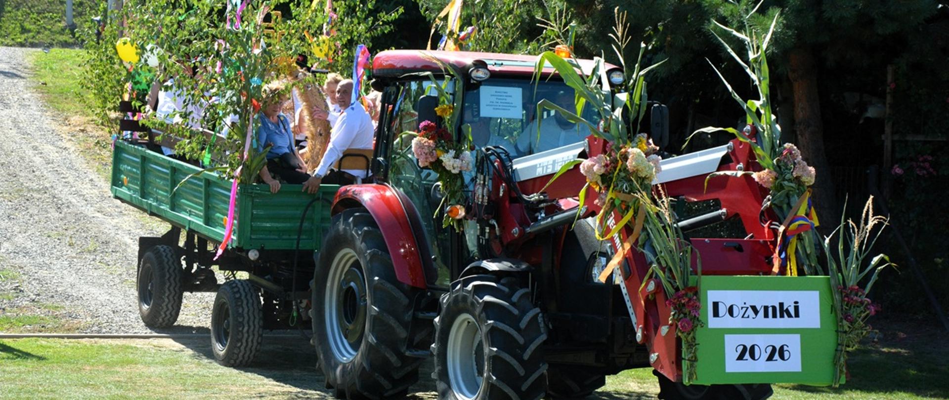 Na zdjęciu znajduje się udekorowany ciągnik z przyczepą, ozdobiony kwiatami, gałęziami, zbożem i kolorowymi wstążkami. Na przyczepie siedzi kilka osób. Z przodu pojazdu umieszczono tablicę z napisem „Dożynki 2026”. W tle widać drzewa, trawnik i zabudowania.