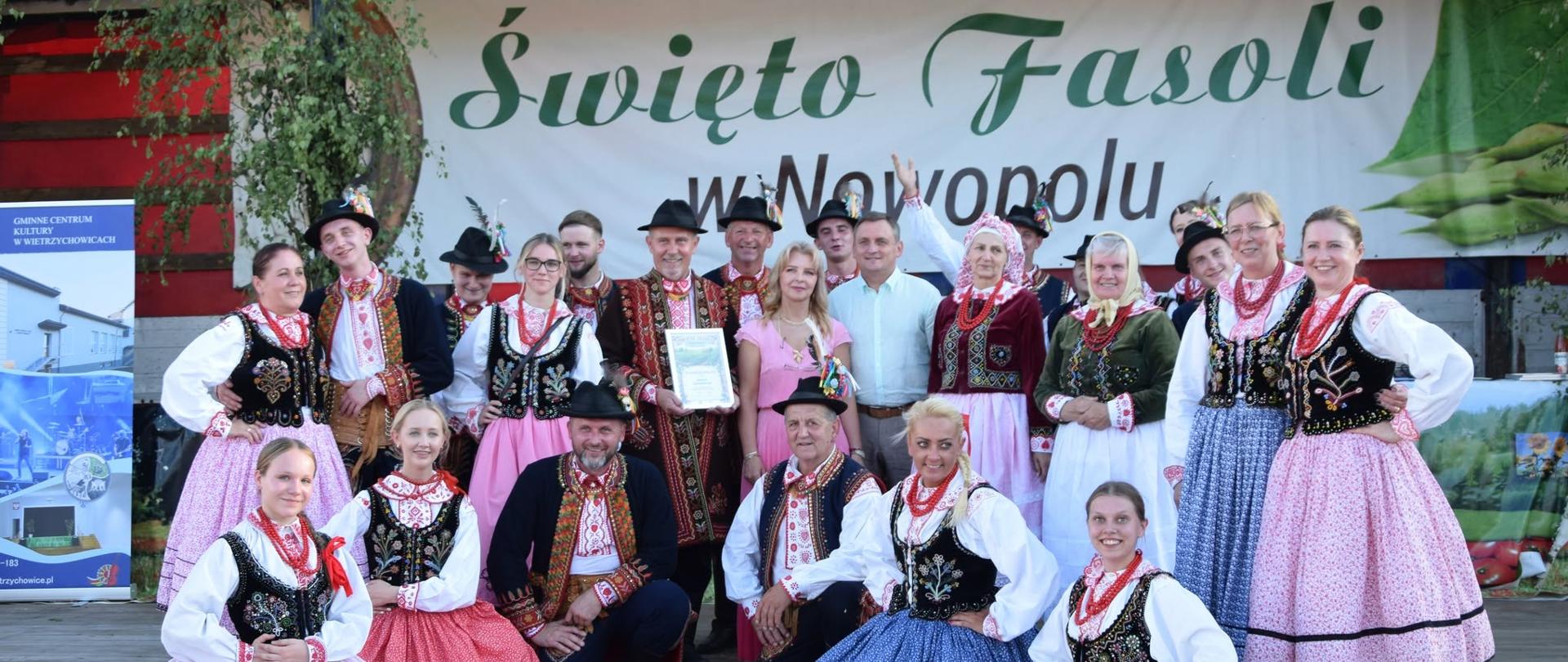 Grupa tancerzy i członków zespołu ludowego pozuje wspólnie na scenie w tradycyjnych, kolorowych strojach regionalnych. W tle widoczny jest baner „Święto Fasoli w Nowopolu”.