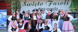 Grupa tancerzy i członków zespołu ludowego pozuje wspólnie na scenie w tradycyjnych, kolorowych strojach regionalnych. W tle widoczny jest baner „Święto Fasoli w Nowopolu”.